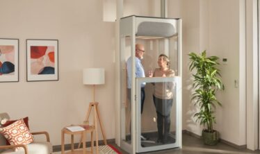 Starší pár v domácím výtahu Stannah Uplifts v prosvětleném obývacím pokoji – vyniká tady kompaktní design i přehledné panely výtahu