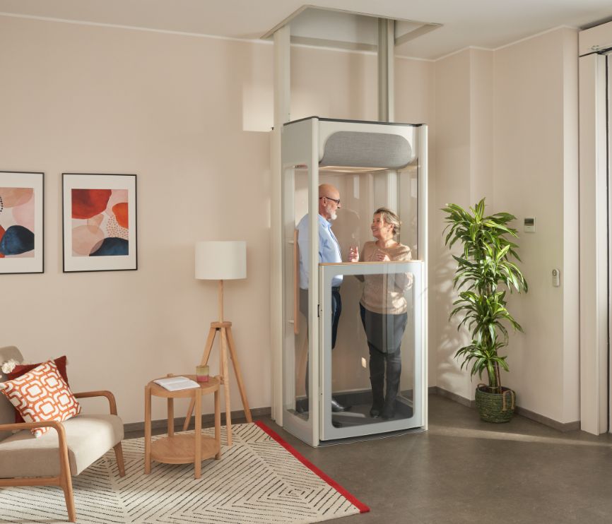 Starší pár v domácím výtahu Stannah Uplifts v prosvětleném obývacím pokoji – vyniká tady kompaktní design i přehledné panely výtahu